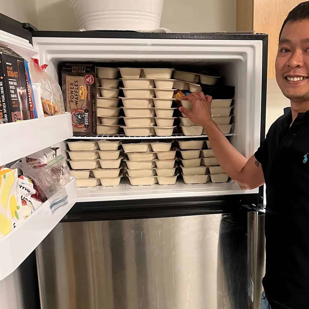 Aly Tran, Food For Free's Logistics Manager, posing next to a freezer full of Heat-n-Eats meals at the Epiphany School in Dorchester.